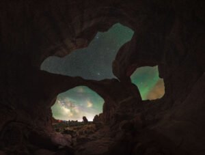 Vista desde el interior de una cueva con dos aberturas que enmarcan la Vía Láctea y auroras boreales verdes en el cielo nocturno estrellado.
