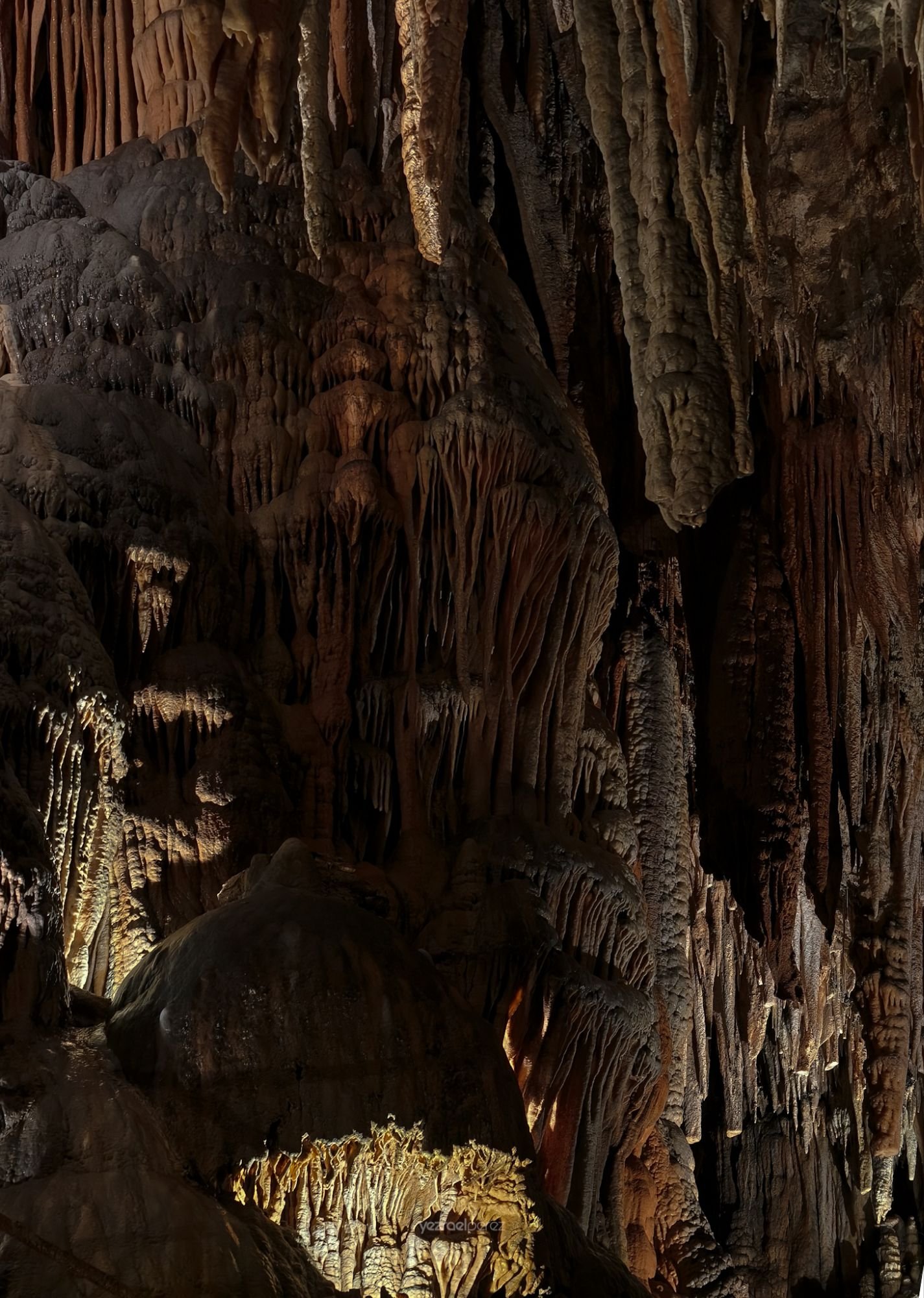 Interior de cueva con estalactitas y estalagmitas iluminadas en tonos dorados y marrones, formaciones calcáreas colgantes del techo y columnas de roca en el suelo.