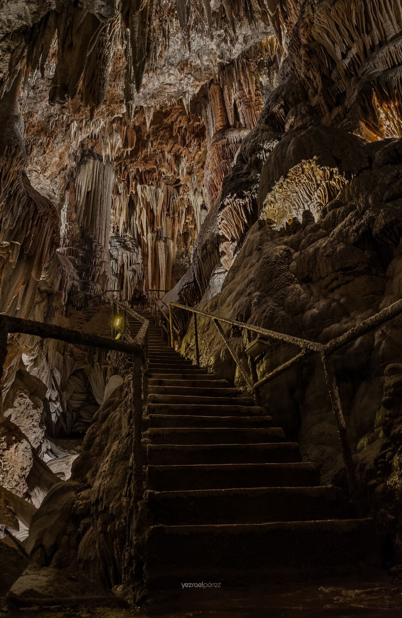 Escaleras de piedra descendentes en caverna con estalactitas y estalagmitas iluminadas, barandilla metálica a los lados