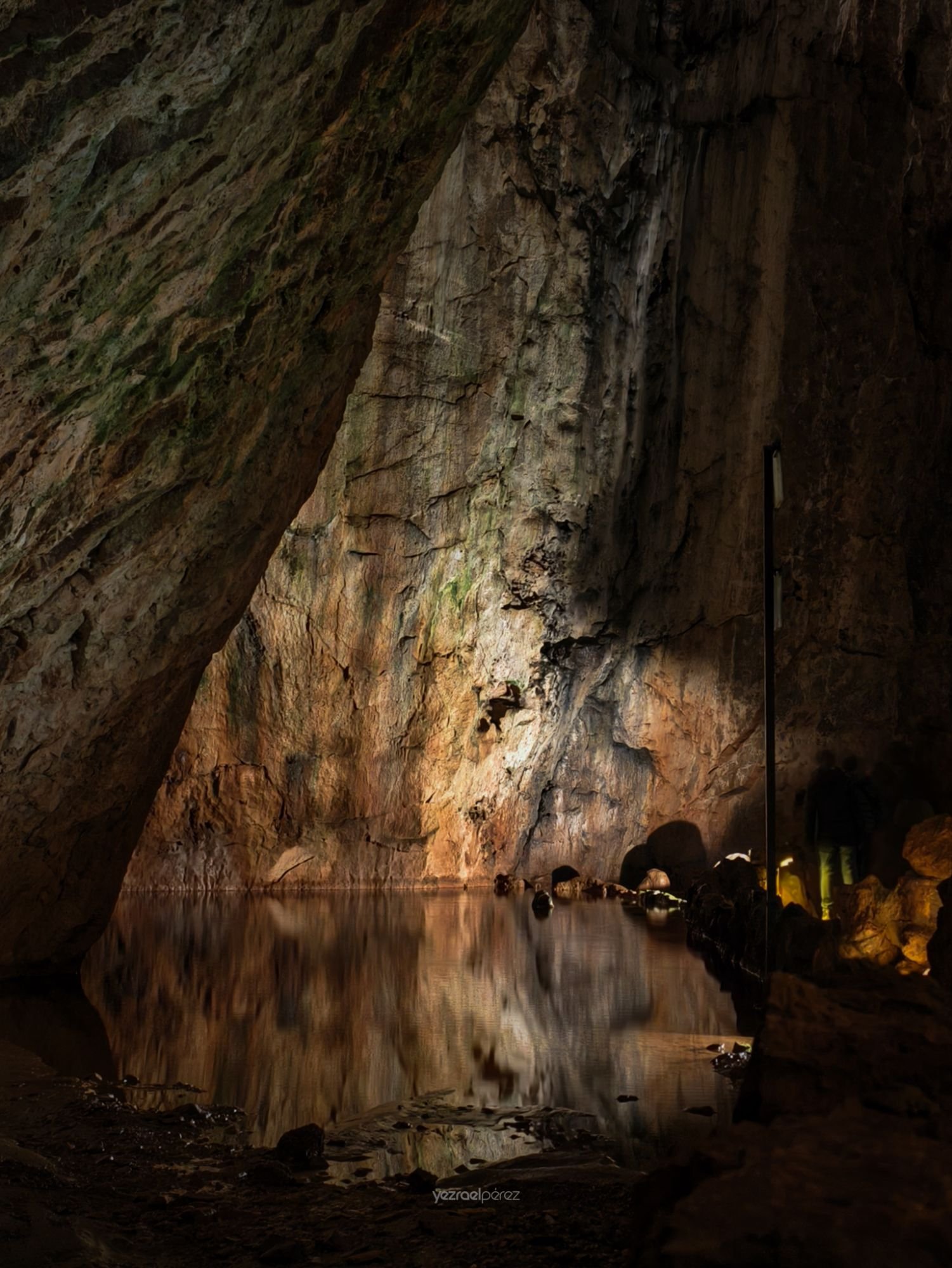 Cueva subterránea con lago subterráneo reflejando paredes rocosas iluminadas, visitantes en la orilla derecha