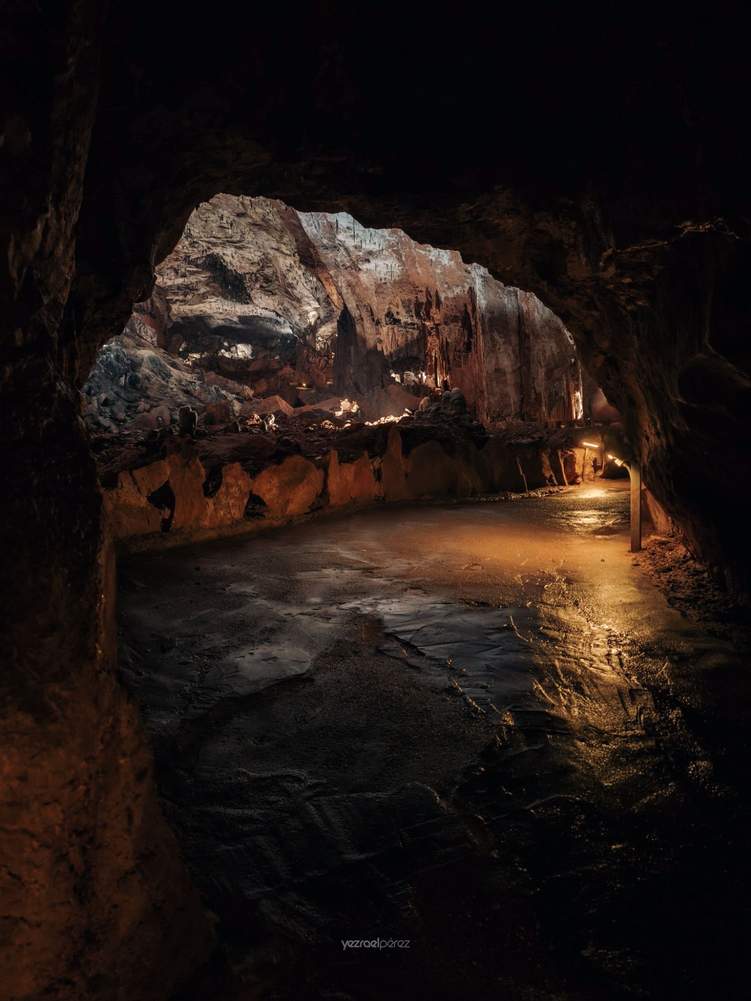 Interior de cueva iluminada con entrada natural de luz, pasarela de madera, formaciones rocosas y reflejo en agua