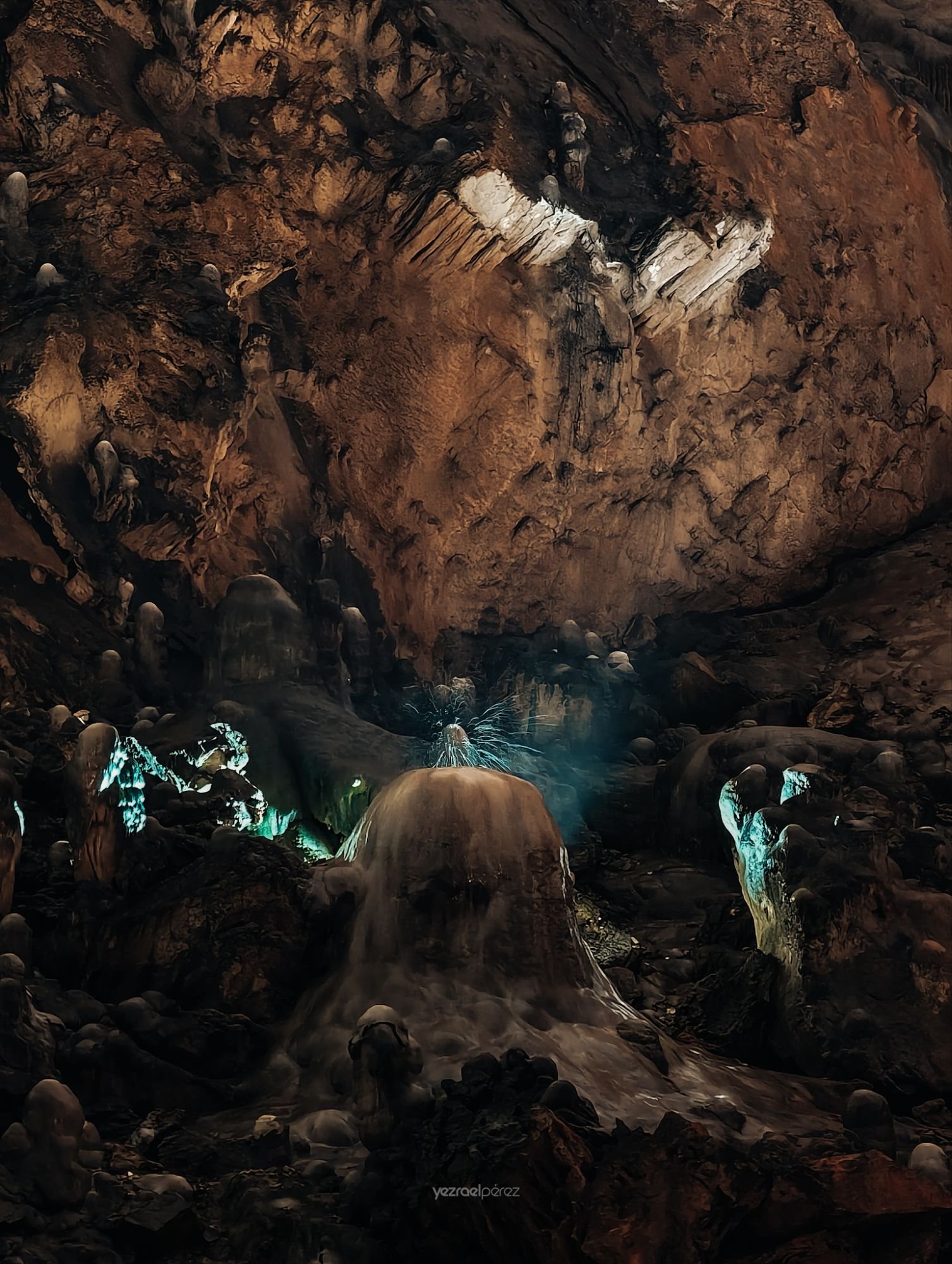 Cueva subterránea con formaciones de estalactitas y estalagmitas iluminadas en azul turquesa, agua cristalina refleja la luz natural que penetra desde la abertura superior