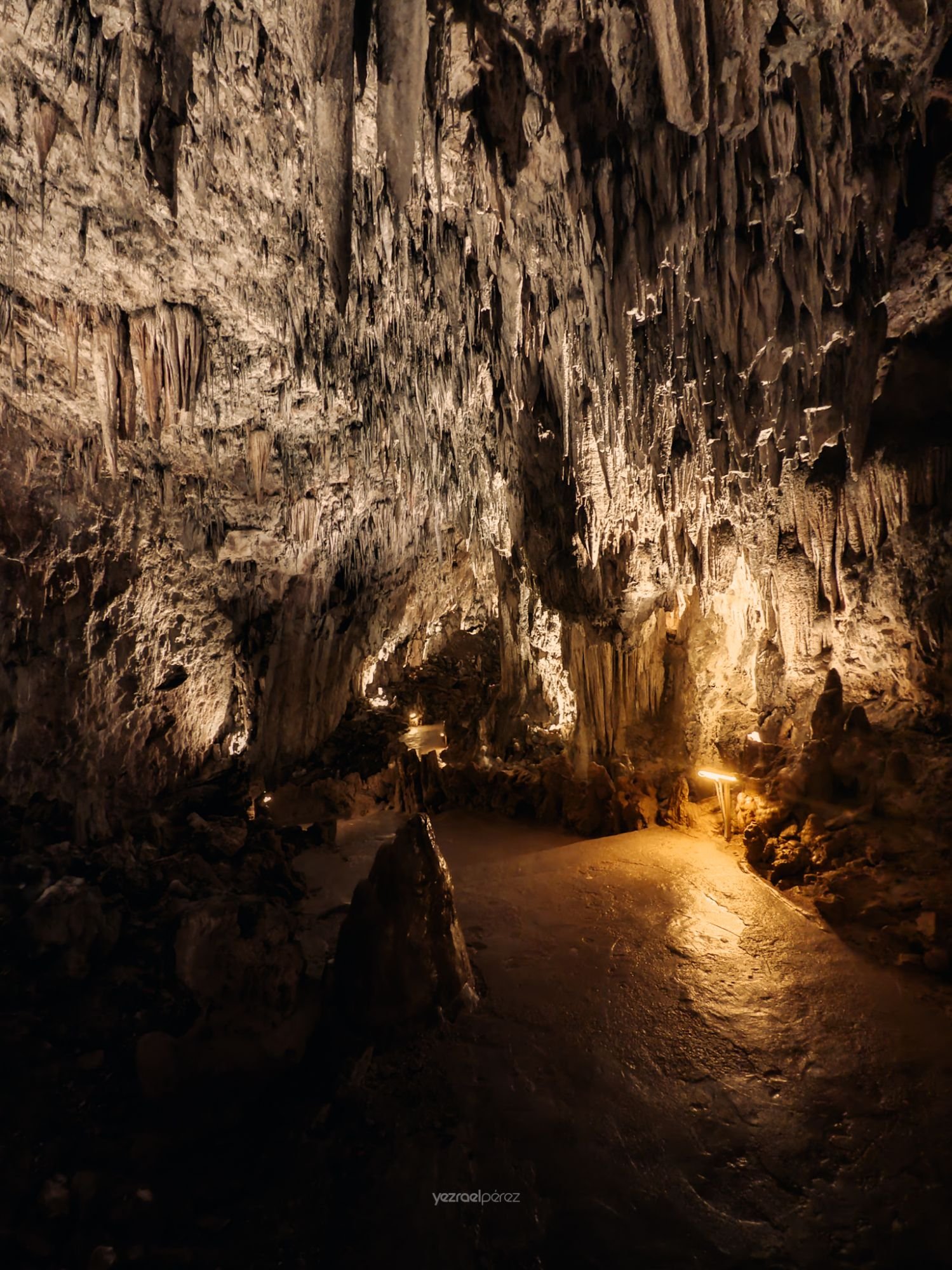 Interior de cueva subterránea con estalactitas y estalagmitas iluminadas, formaciones calcáreas colgantes desde el techo y suelo rocoso