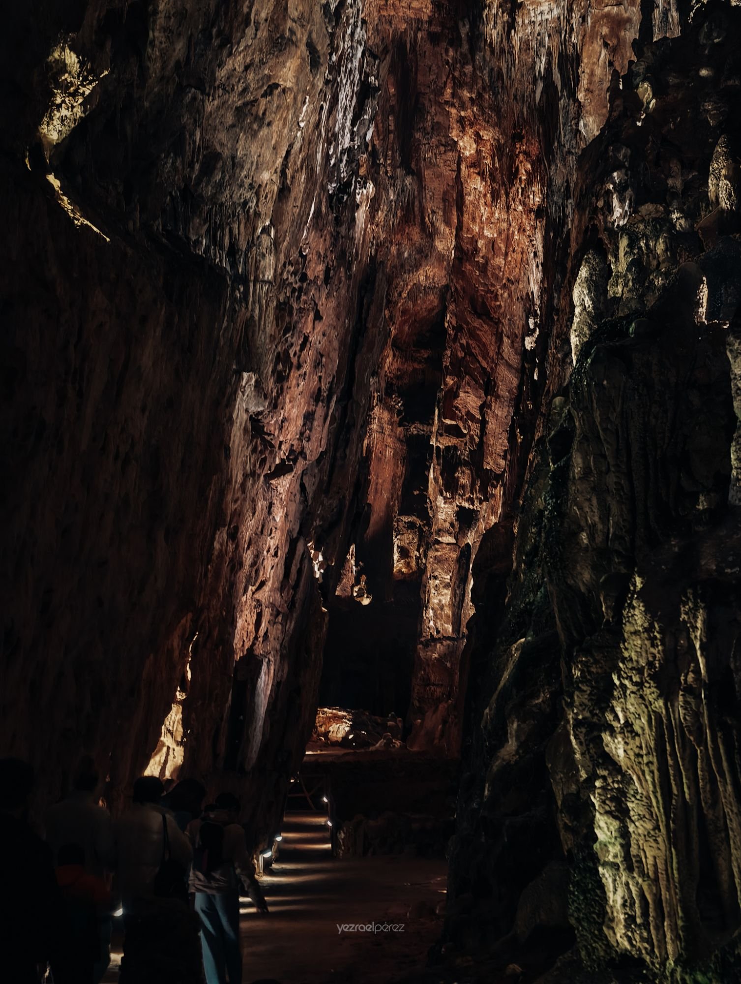 Pasaje subterráneo estrecho entre paredes de roca oscura con tonos rojizos y verdosos, iluminado por luz natural que filtra desde arriba creando sombras dramáticas.