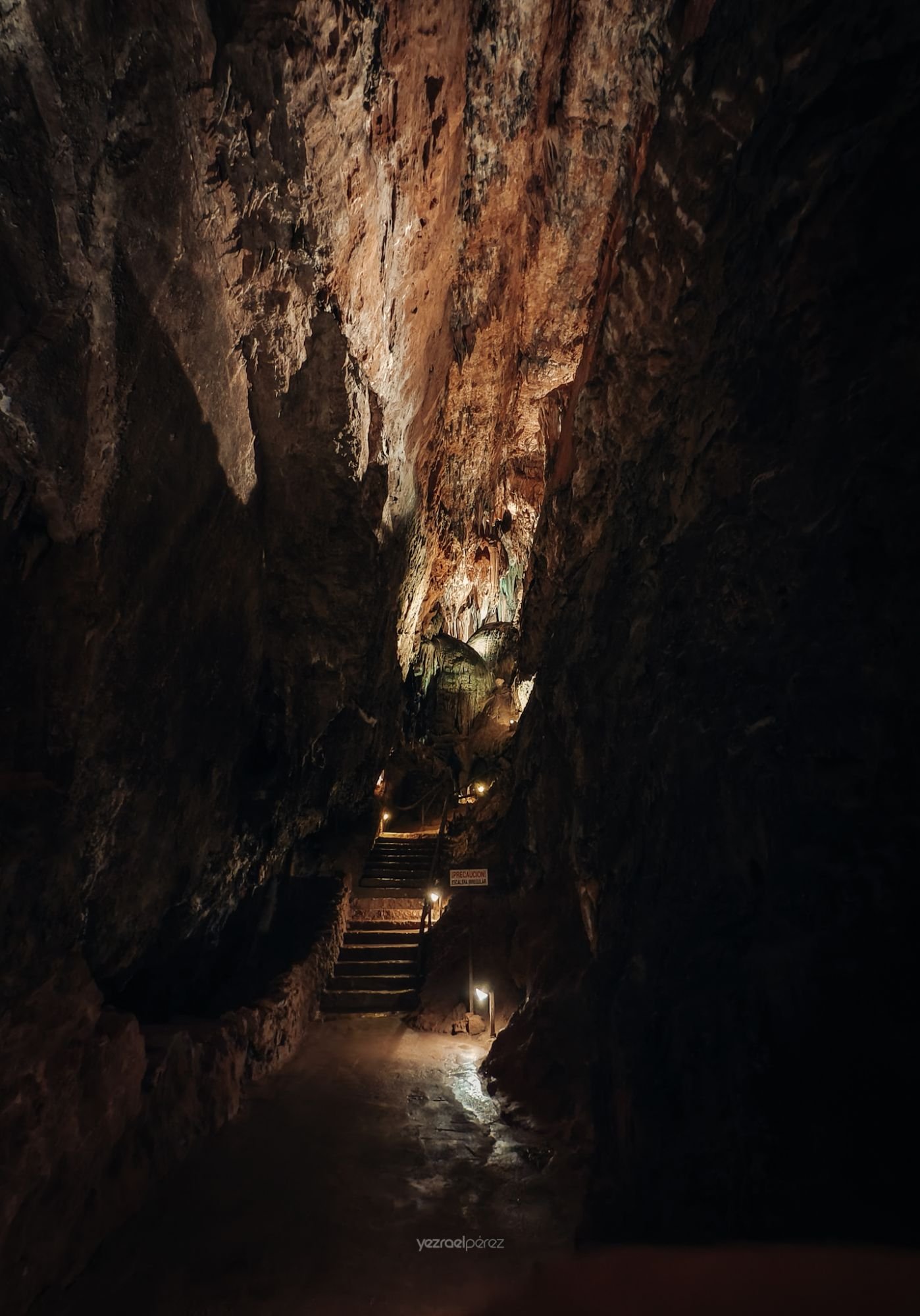 Interior de cueva subterránea iluminada con escaleras de piedra, paredes rocosas oxidadas en tonos ocres y negros, luz artificial revelando formaciones geológicas.