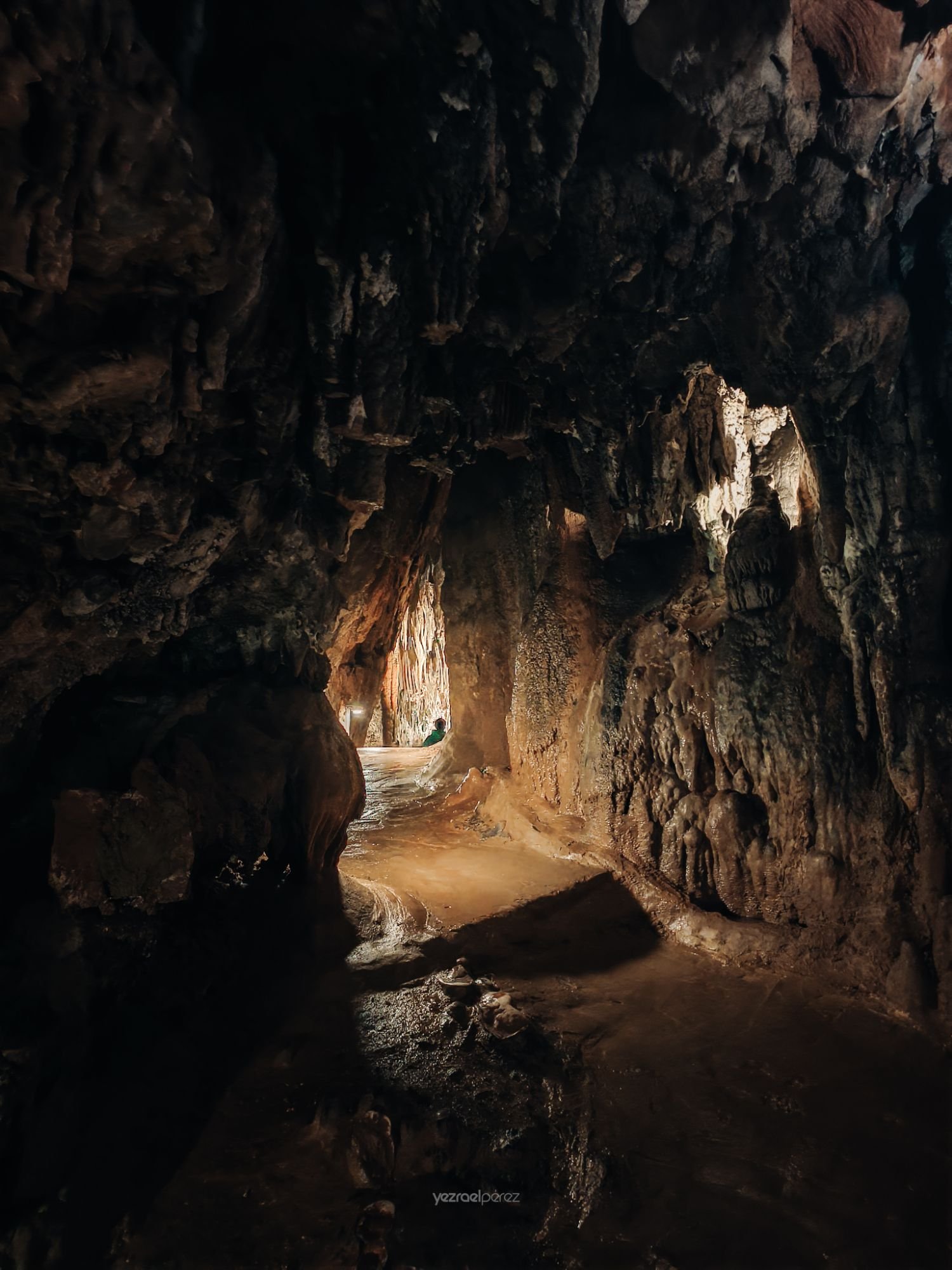 Interior de cueva con paredes de roca oscura, luz natural filtrándose por abertura al fondo iluminando arena y formaciones rocosas