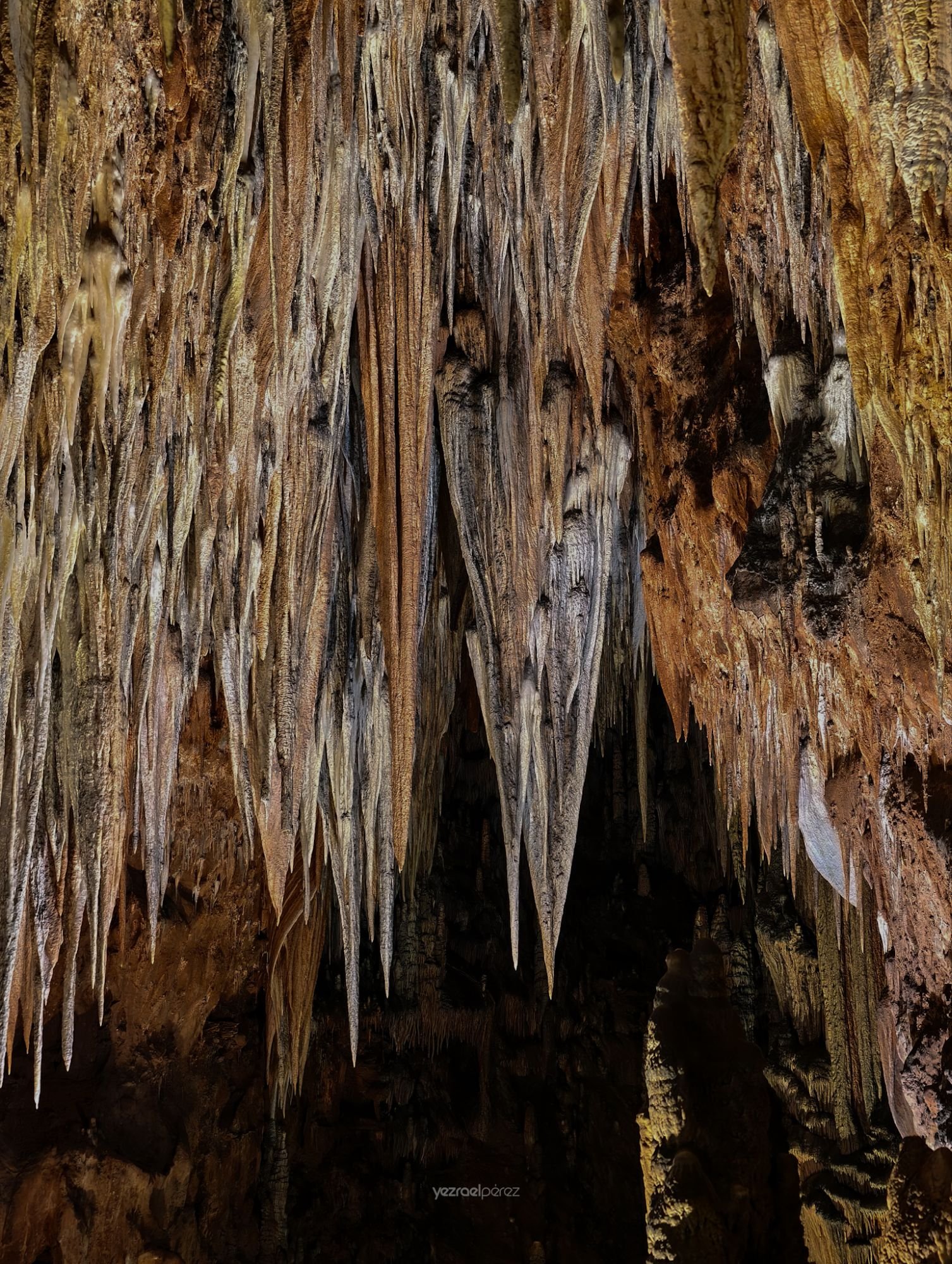 Estalactitas de piedra caliza colgando del techo de una cueva, con tonos marrones, grises y blancos, iluminadas por luz natural que revela la textura erosionada de la roca