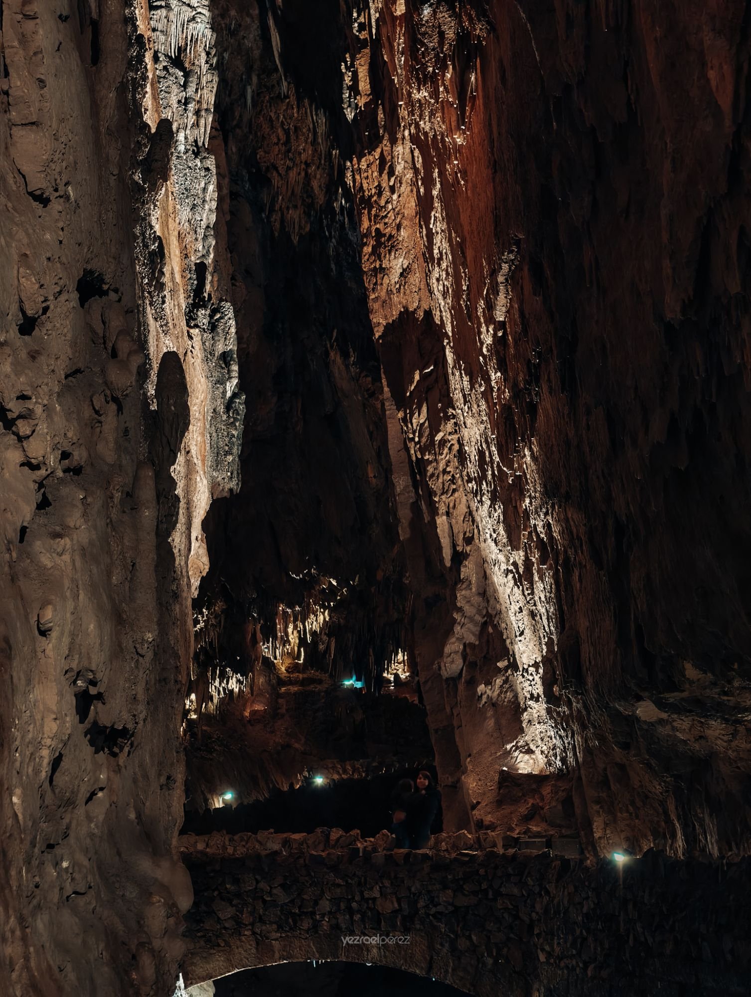 Interior de caverna subterránea con formaciones de estalactitas blancas iluminadas, paredes de roca oscura y luces azules en el suelo.