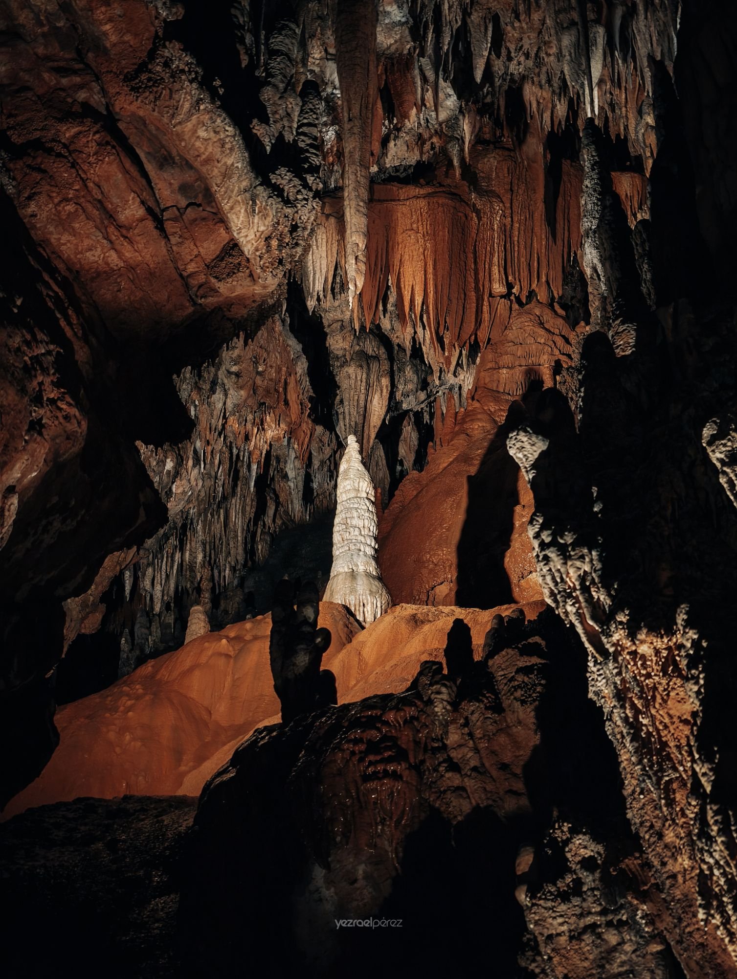 Caverna subterránea con estalagmitas y estalactitas de colores ocre y blanco, iluminadas dramáticamente contra la oscuridad de la cueva.