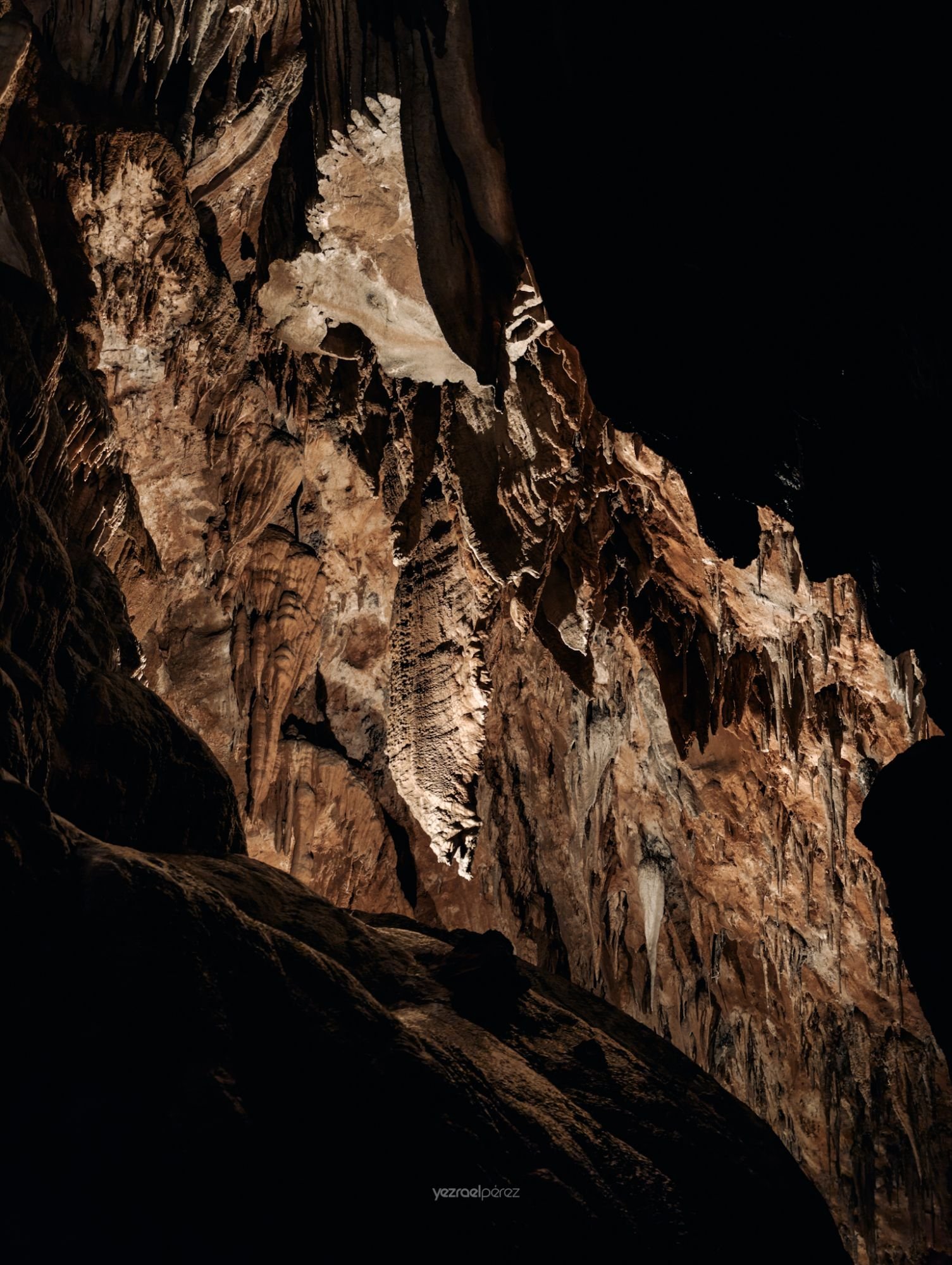 Interior de cueva con formaciones de estalactitas y estalagmitas iluminadas en tonos ocres y blancos, fotografiado desde la oscuridad.