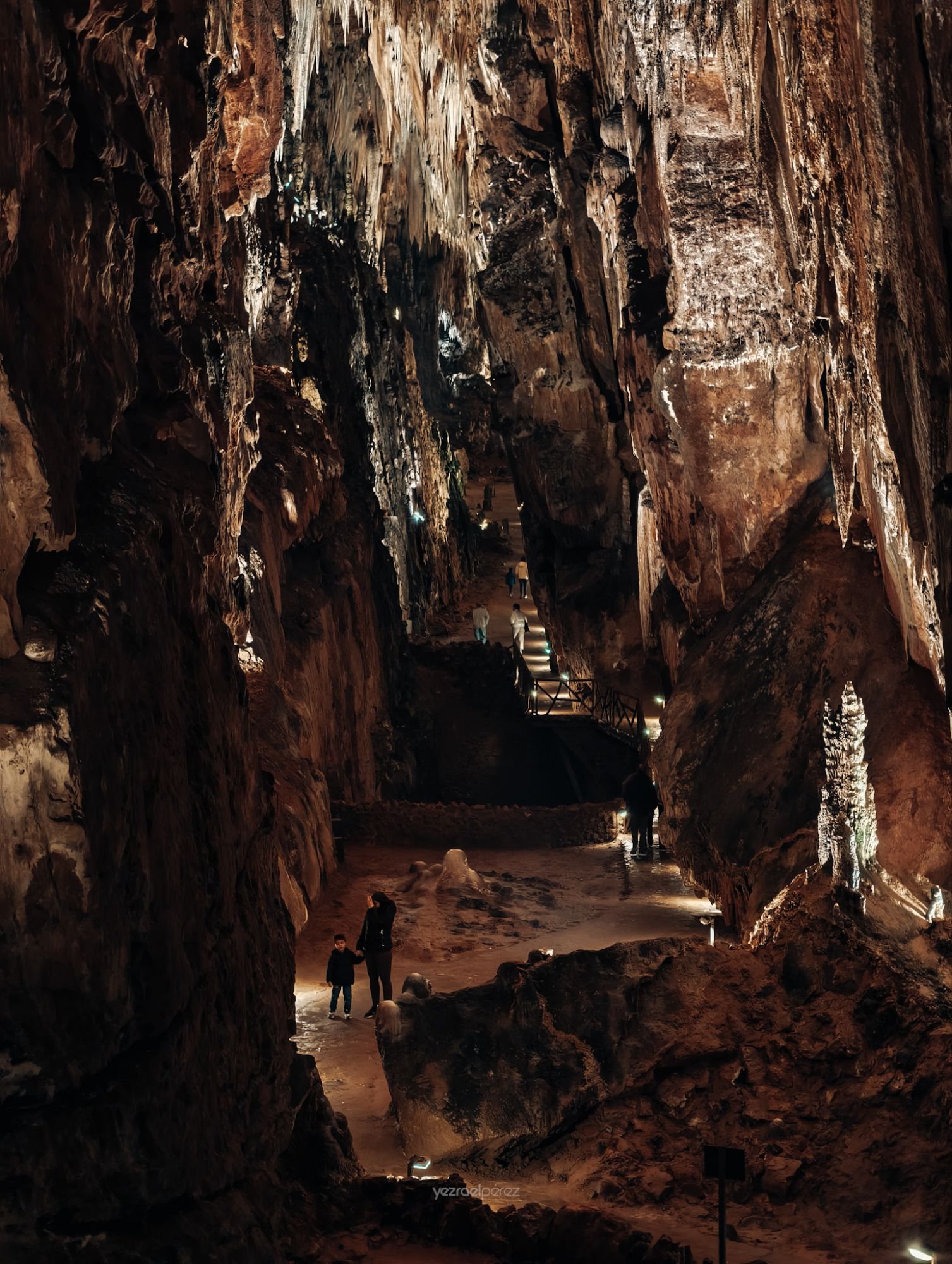Interior de cueva con paredes rocosas de tonos marrones y blancos. Visitantes caminan por el suelo de arena roja iluminados por luz natural que filtra desde arriba.