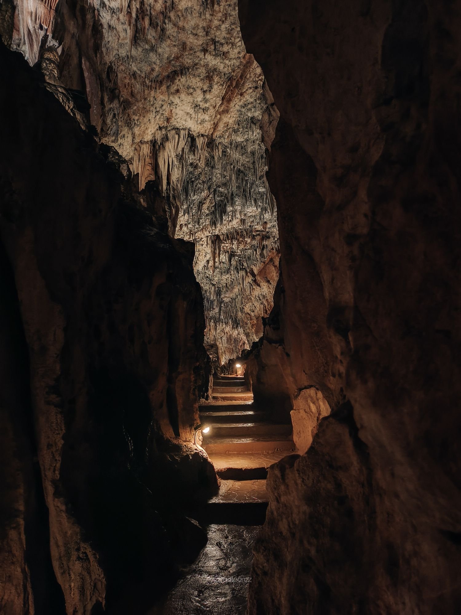 Interior de caverna iluminada con estalactitas calcáreas colgantes, pasillo de piedra con escaleras y luz cálida al fondo