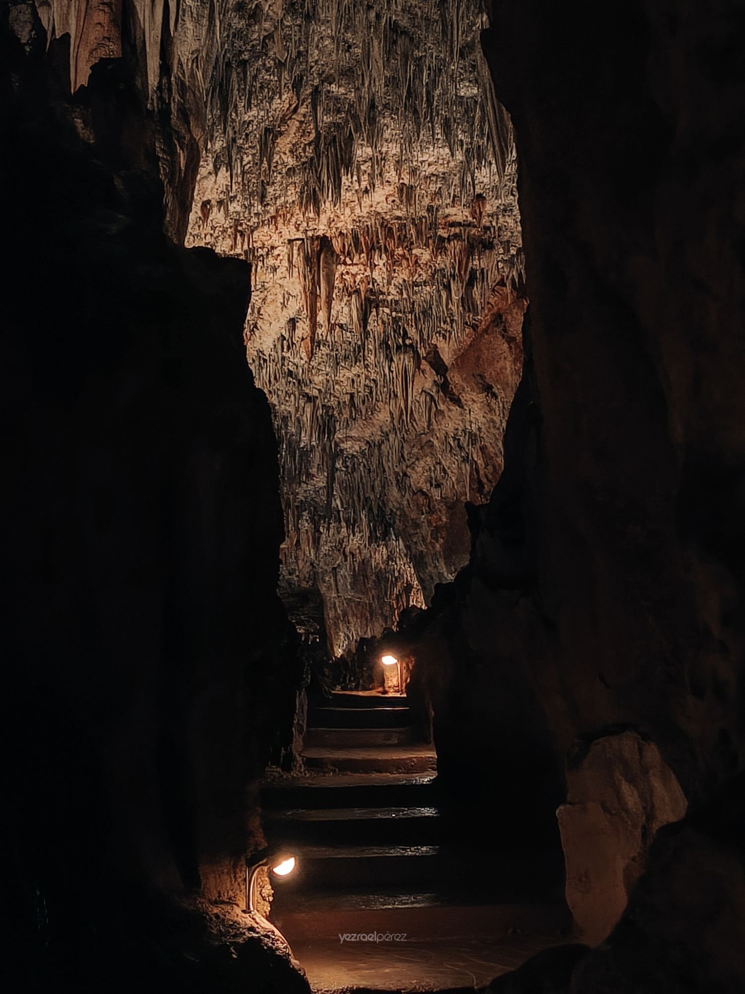 Interior de cueva con estalactitas iluminadas por luces cálidas, escaleras de piedra descendiendo hacia una cámara subterránea