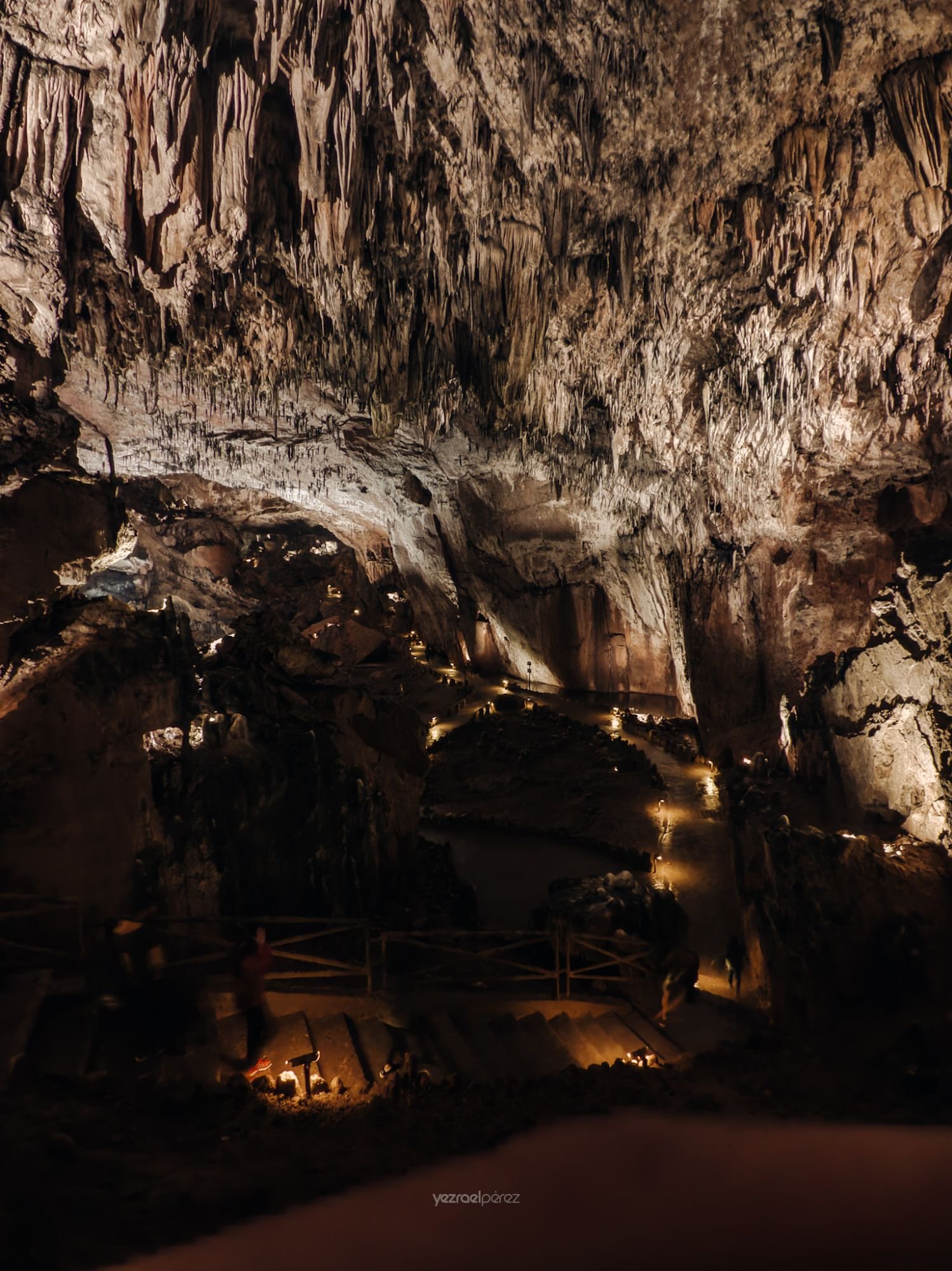 Caverna subterránea iluminada con estalactitas en el techo, pasarela de madera y luces cálidas reflejadas en agua oscura