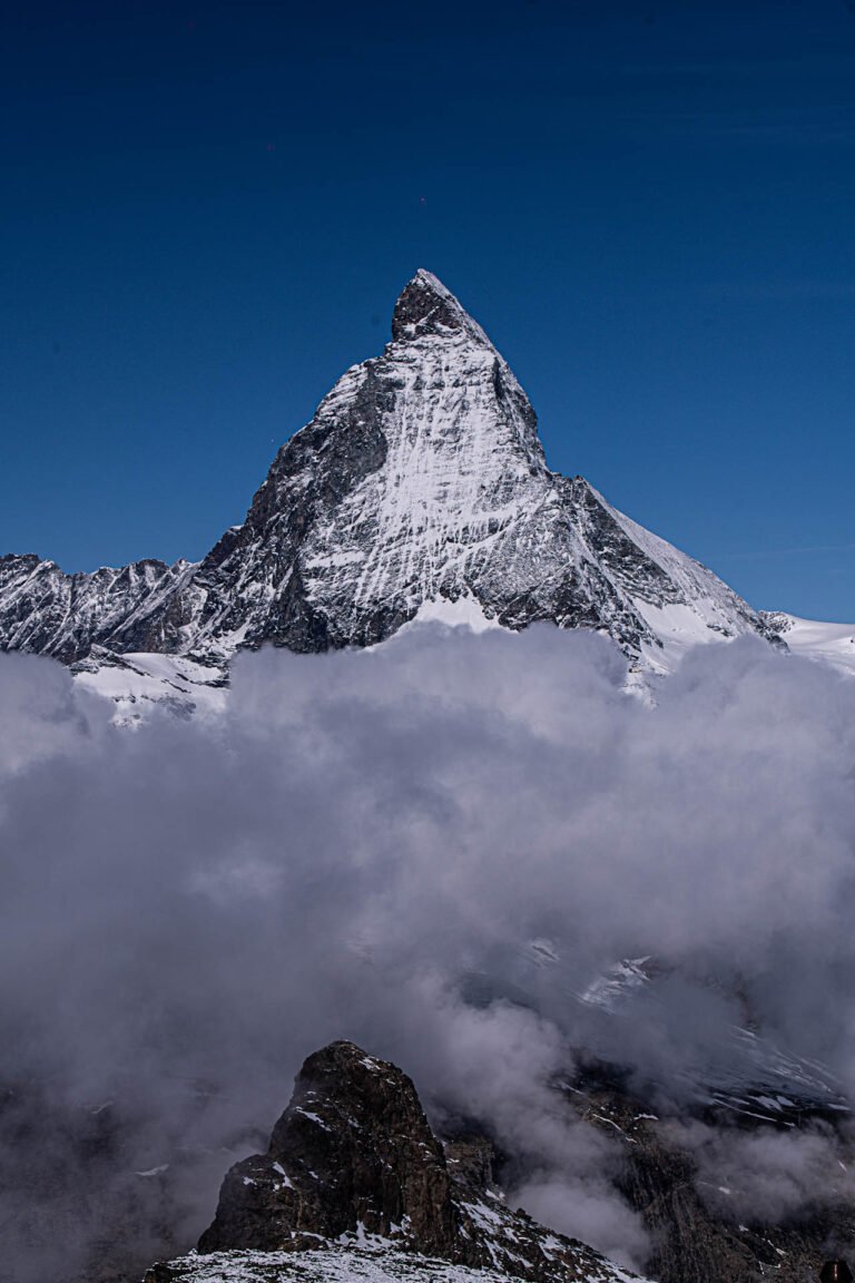 Matterhorn nevado emergiendo entre nubes densas bajo cielo azul despejado, su pico pyramidal cubierto de nieve y roca oscura